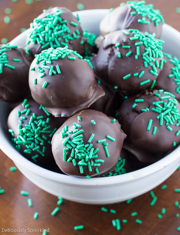 A white bowl filled with mint oreo truffles is surrounded by green sprinkles. 