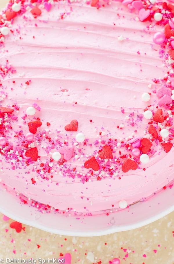 A pink frosted cake is presented on a white cake stand and topped with pink, white, and red sprinkles.