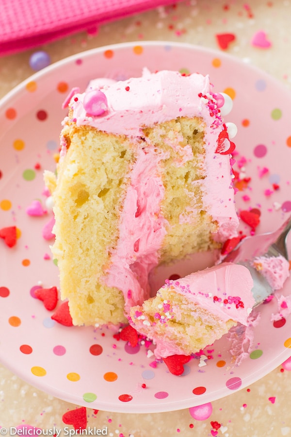 A fork is removing a bite sized portion of vanilla cake with frosting from a pink plate.