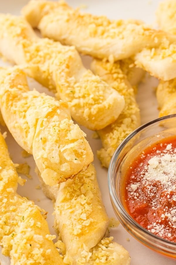 A few breadsticks are placed around a cup of marinara sauce.