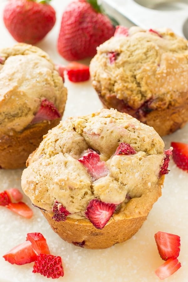 Three strawberry muffins are placed next to berries on a white surface.
