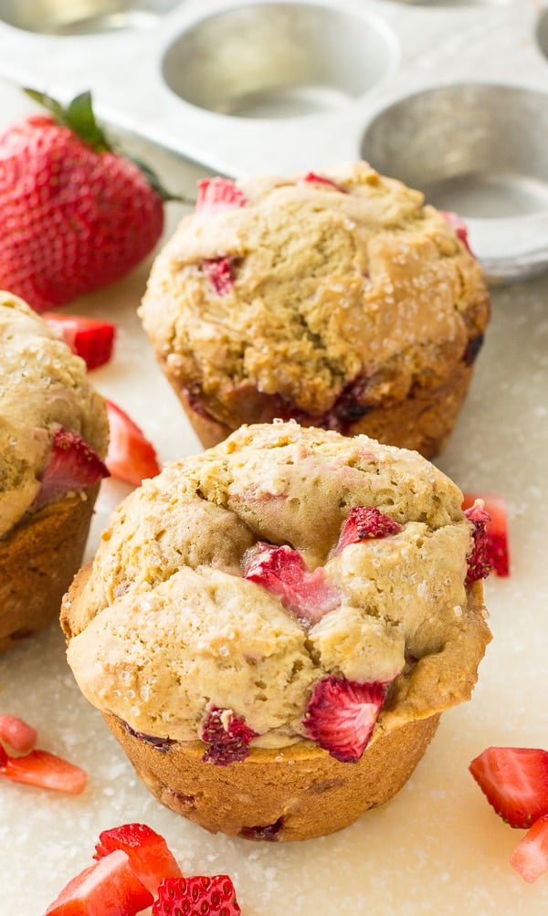 Strawberry muffins are placed next to additional berries and an empty baking tin on a white surface.