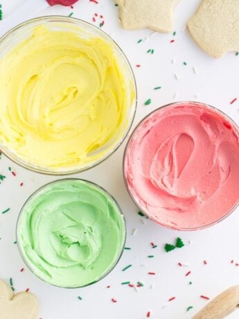 Best sugar cookie frosting in bowls colored yellow, green, and pink with sprinkles and cookies scattered around.