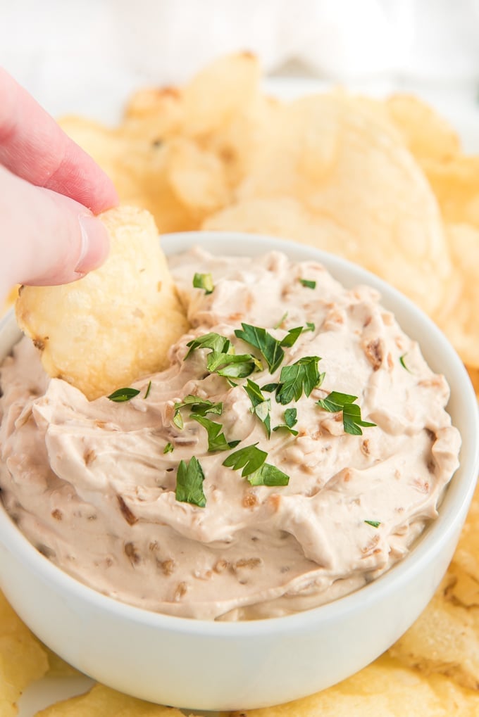 A potato chip is being dipped into a small white bowl filled with dip.