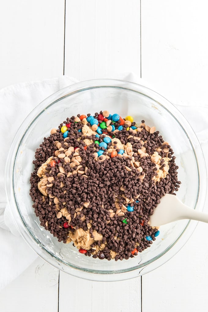 Chocolate chips and M&Ms have been added to a glass bowl filled with cookie dough. 