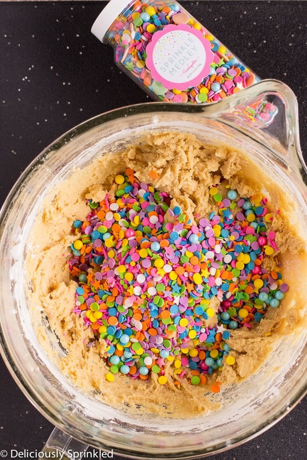 Cookie dough is being mixed with sprinkles in a large glass mixing bowl.