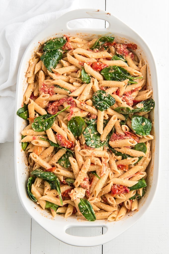 Pasta is stirred with baby spinach in a white baking dish.