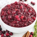A bowl of cranberry sauce with orange juice in a bowl on the table with cinnamon sticks, rosemary and fresh cranberries around it.