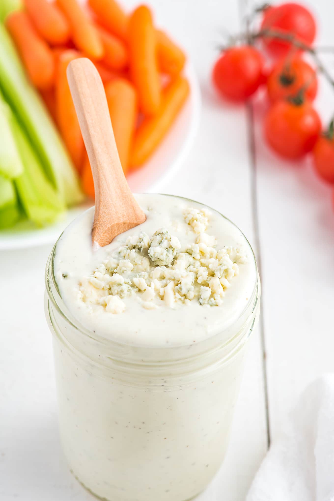 A jar of blue cheese crumbles dressing with a spoon in the jar in front of a platter of veggies.