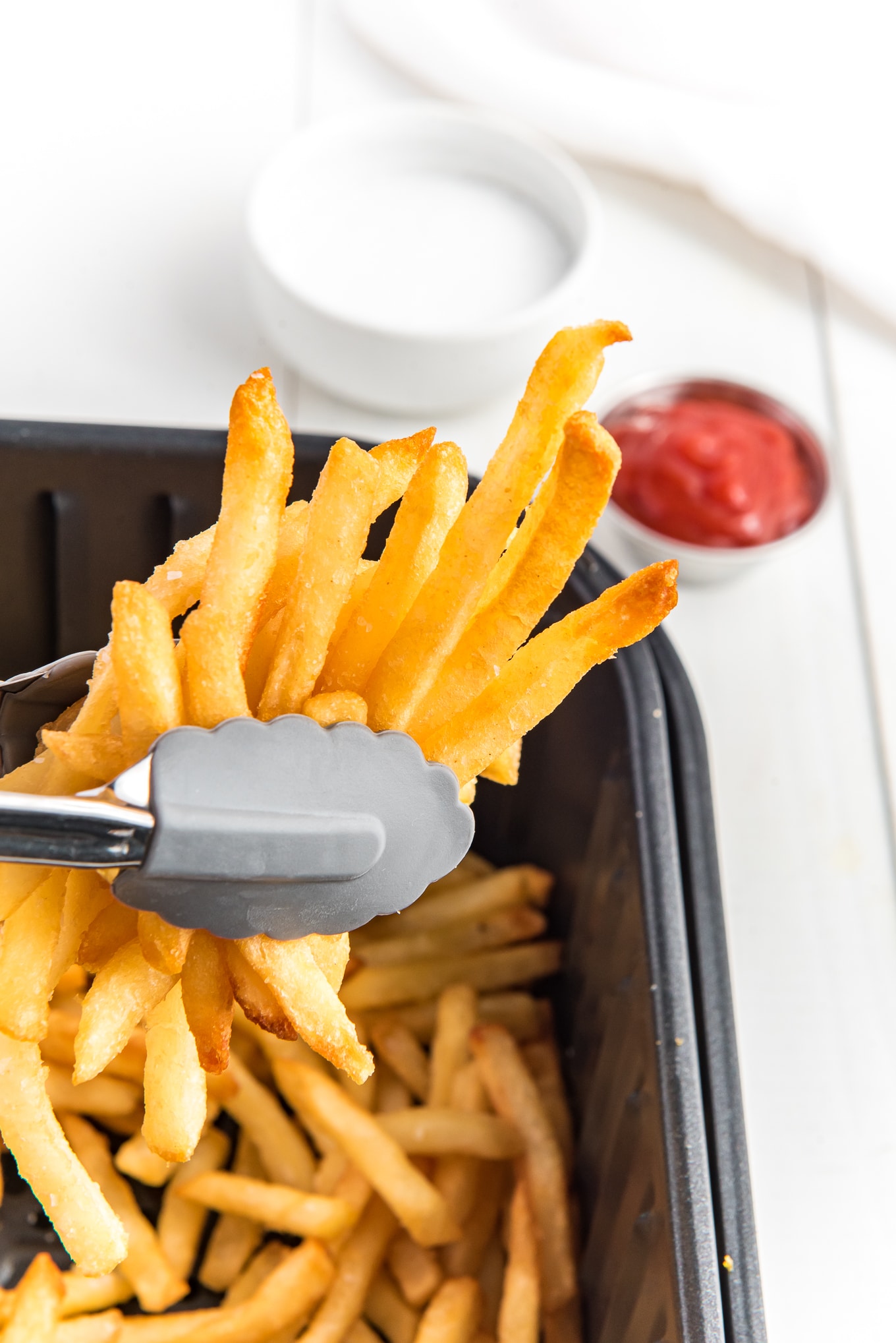 Close up of french fries in kitchen tongs from the air fryer./ Enjoy crispy and delicious frozen French fries in minutes with this easy air fryer recipe. Get that classic crispy texture and savory flavor you love, with the convenience of the air fryer.