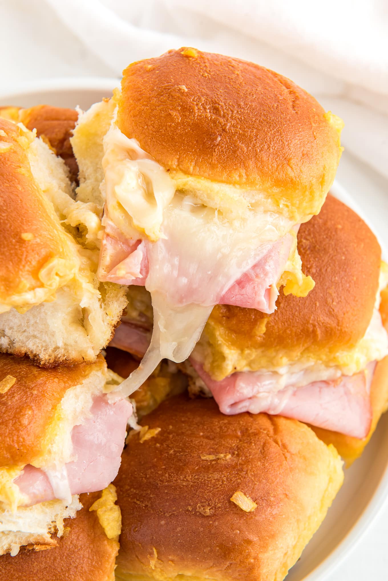 Hawaiian Rolls Sliders with ham and Swiss cheese stacked on a plate./ Delicious ham and cheese sliders on soft and sweet Hawaiian rolls, stacked with melted cheese, juicy ham, and topped with a golden-brown glaze, perfect for a quick dinner or appetizer. 