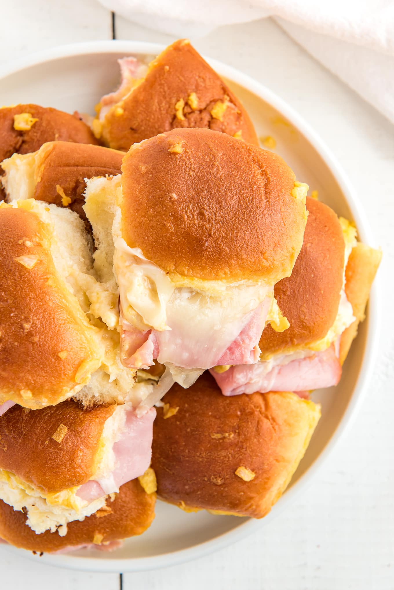 Hawaiian Rolls Sliders with ham and Swiss cheese stacked on a white plate./ Delicious ham and cheese sliders on soft and sweet Hawaiian rolls, stacked with melted cheese, juicy ham, and topped with a golden-brown glaze, perfect for a quick dinner or appetizer. 
