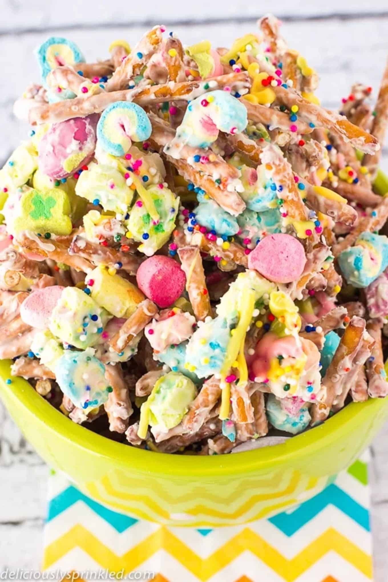 Lucky Charms Party Mix is presented in a green serving bowl. / Lucky Charms marshmallows are tossed together with salty pretzels, M&M candies, and rainbow sprinkles, all coated with sweet white chocolate. 