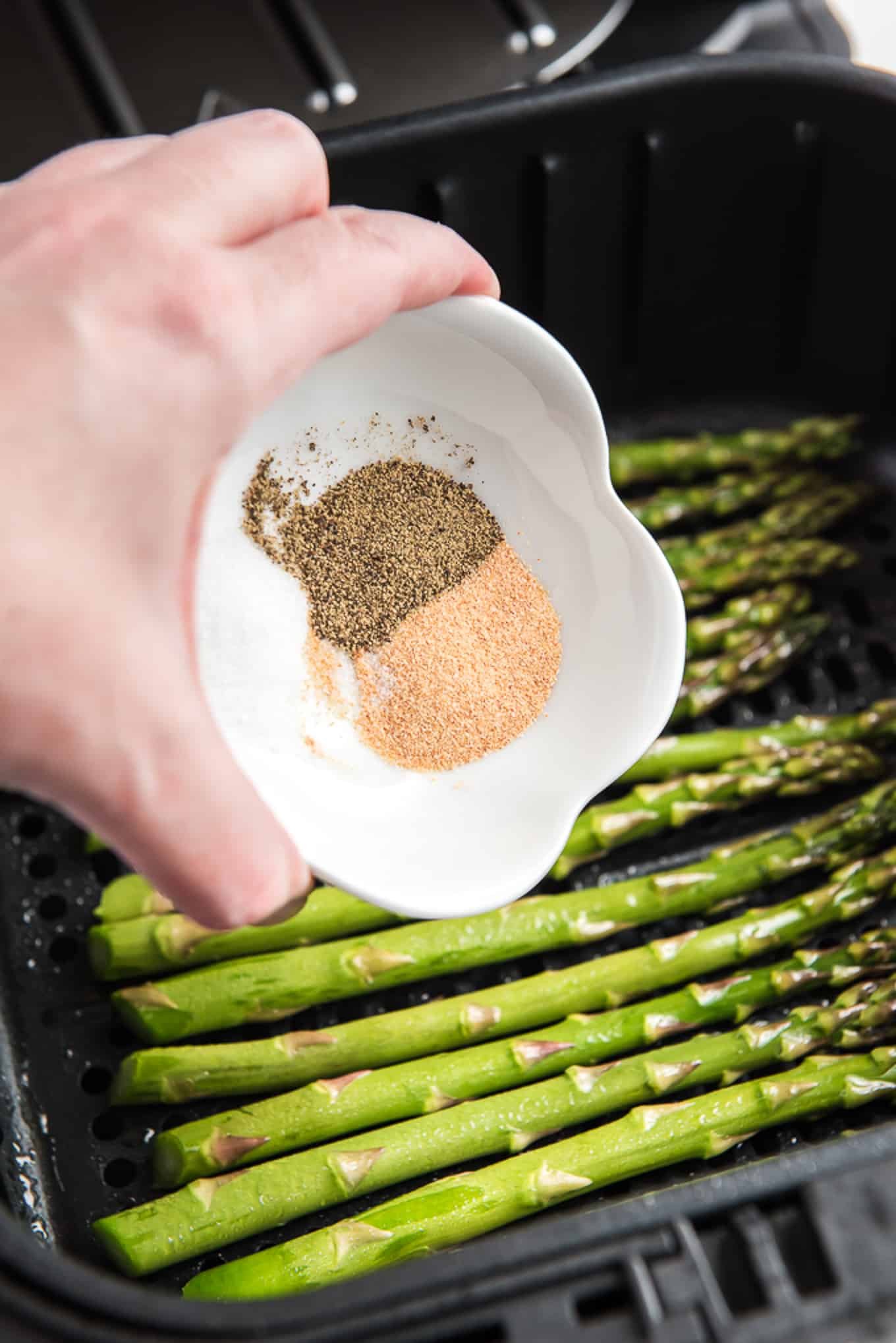 Pouring spices onto the prepared asparagus in the air fryer basket. 