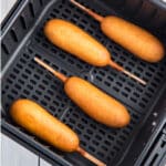 A close-up photo of four corn dogs in an air fryer basket.