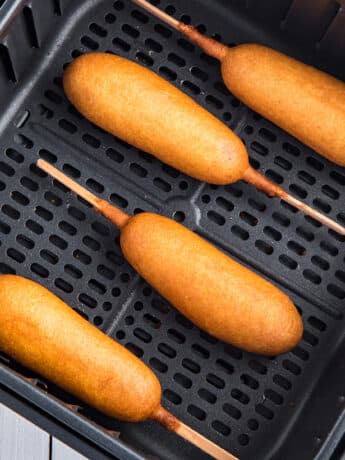 A close-up photo of four corn dogs in an air fryer basket.