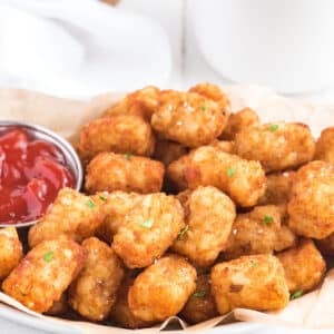A close-up photo of a basket of air fryer tater tots. The tots are golden brown and crispy, and they are piled high on in the basket. There is a small bowl of ketchup on the side of the basket.