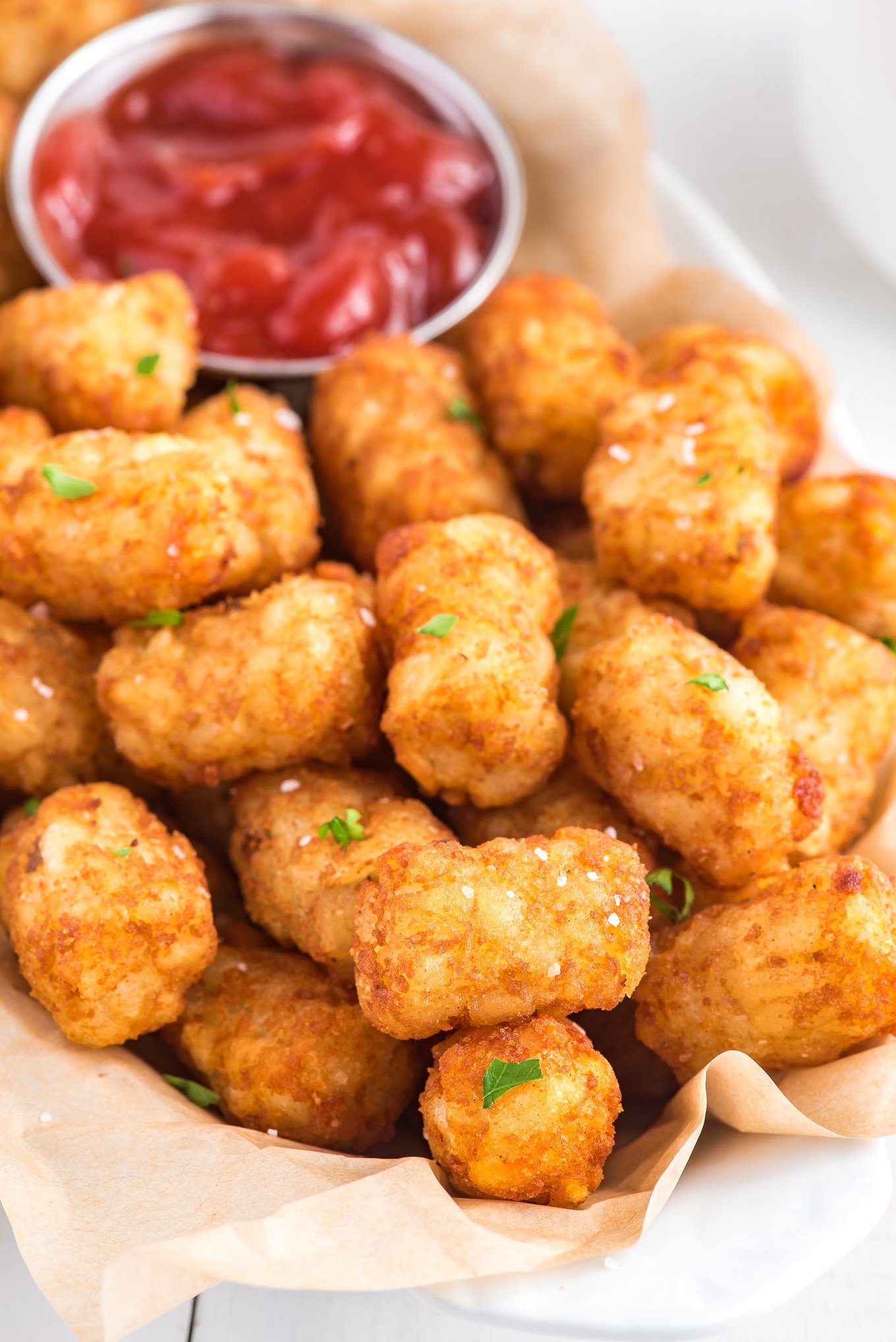 A close-up photo of a basket of air fryer tater tots. The tots are golden brown and crispy, and they are piled high on in the basket. There is a small bowl of ketchup on the side of the basket.