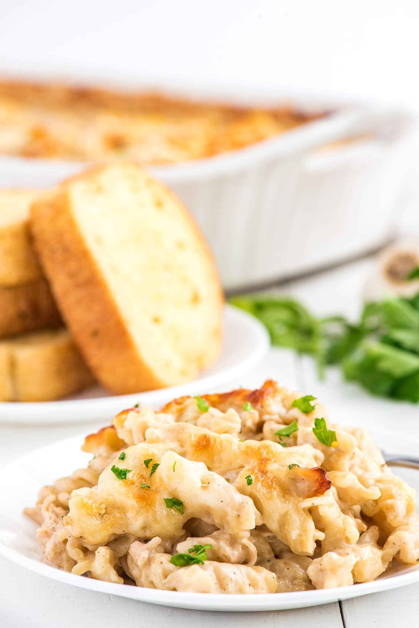 A plate of chicken alfredo bake pasta with parsley on top and a plate of garlic bread in the back.