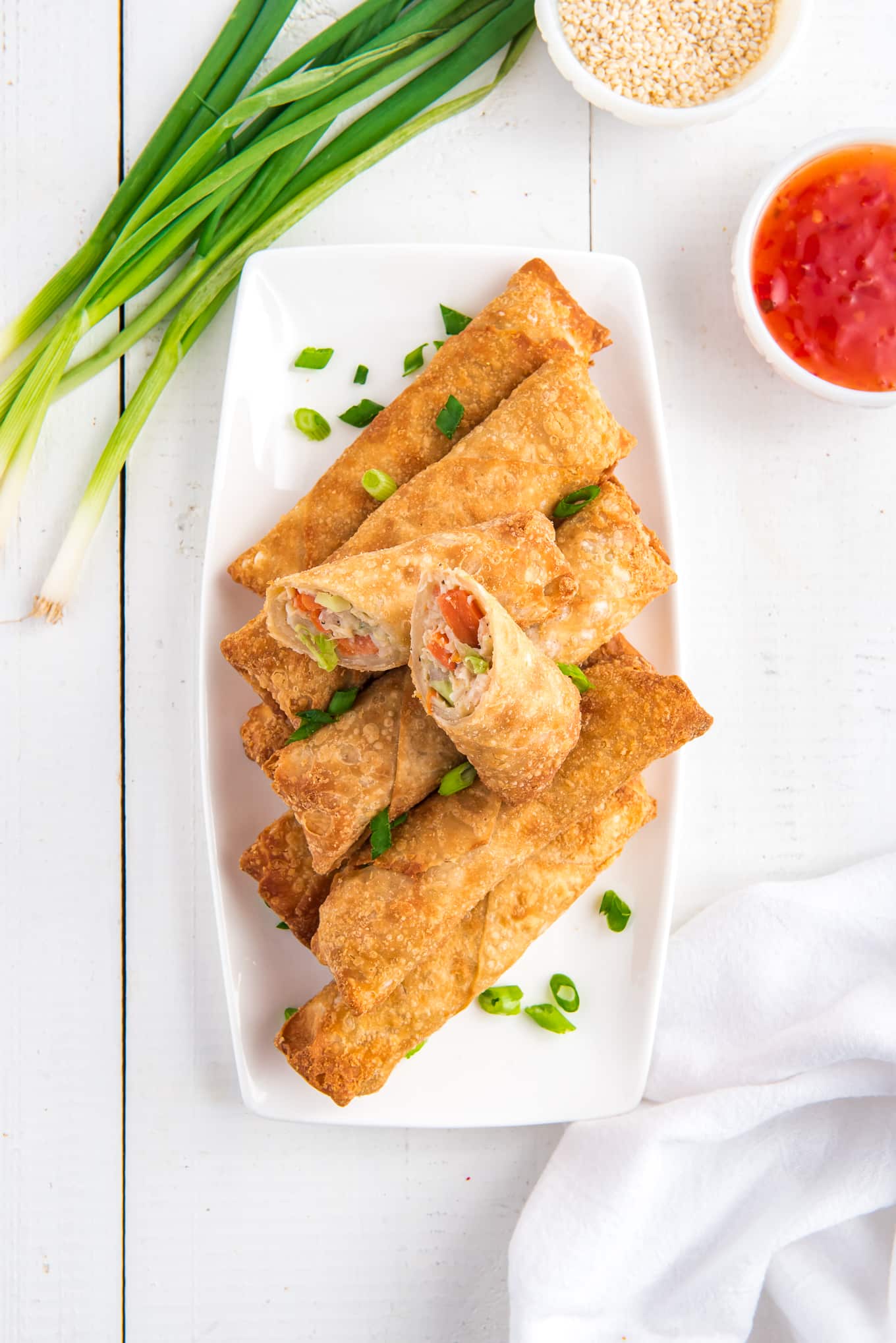 Plate with air fryer egg rolls with a side of sweet chili sauce, a bowl of sesame seeds and green onions for garnish. 