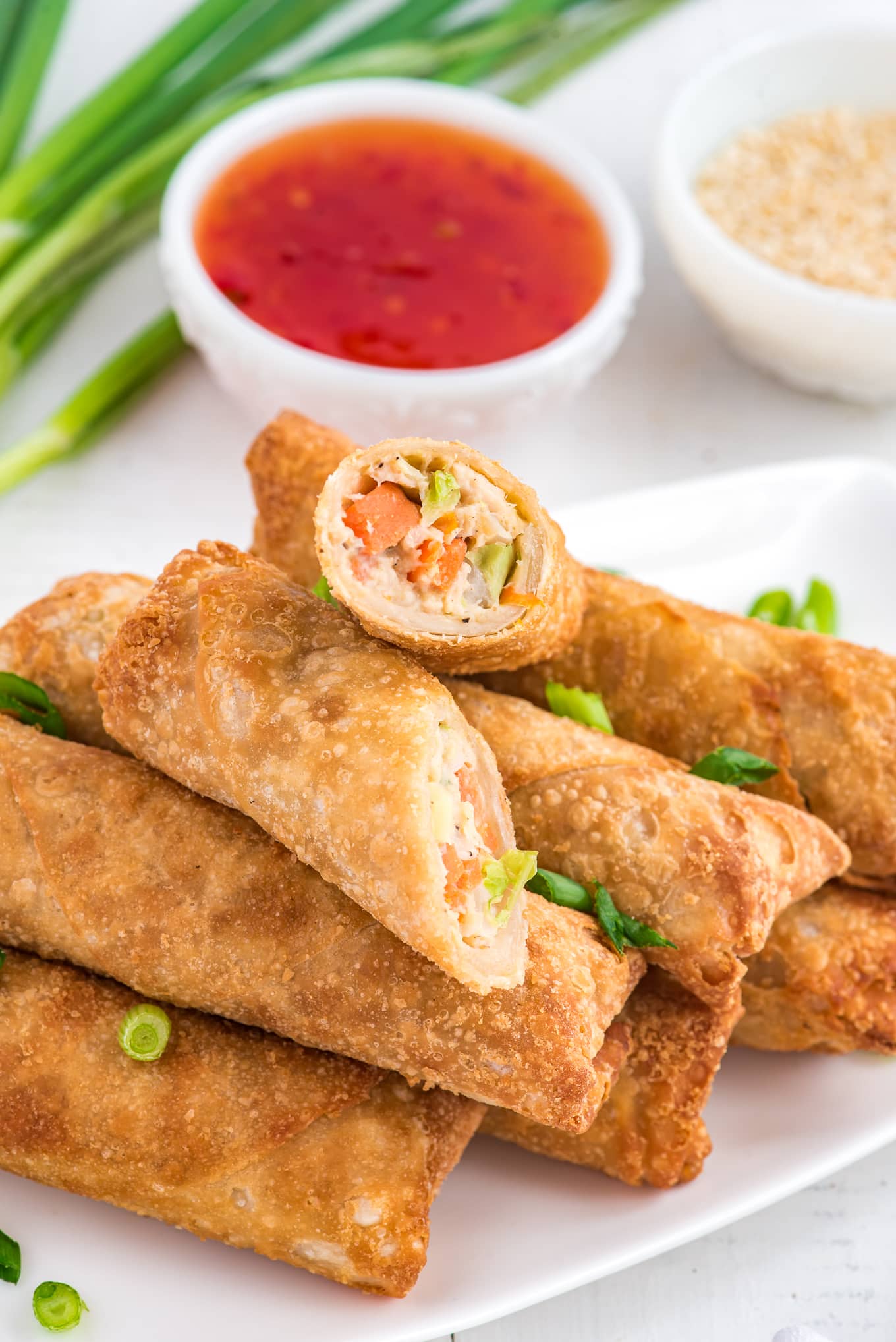 Crispy air fryer egg rolls in a stack with sweet and spicy sauce. One egg roll is cut in half to show texture. Garnished with green onion. 
