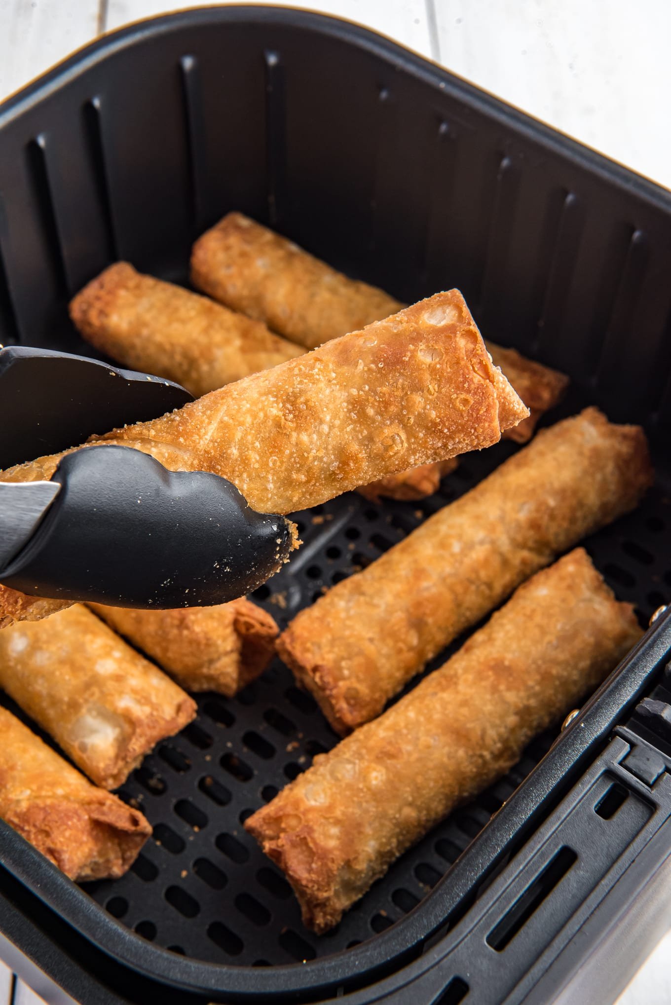 Air fryer egg rolls in the air fryer with a pair of tongs holding and cooked egg roll.