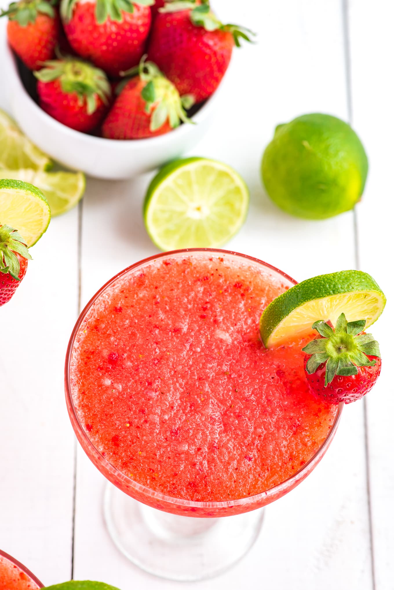 Overhead view of frozen strawberry margarita in a glass with limes and strawberries on the table around it.