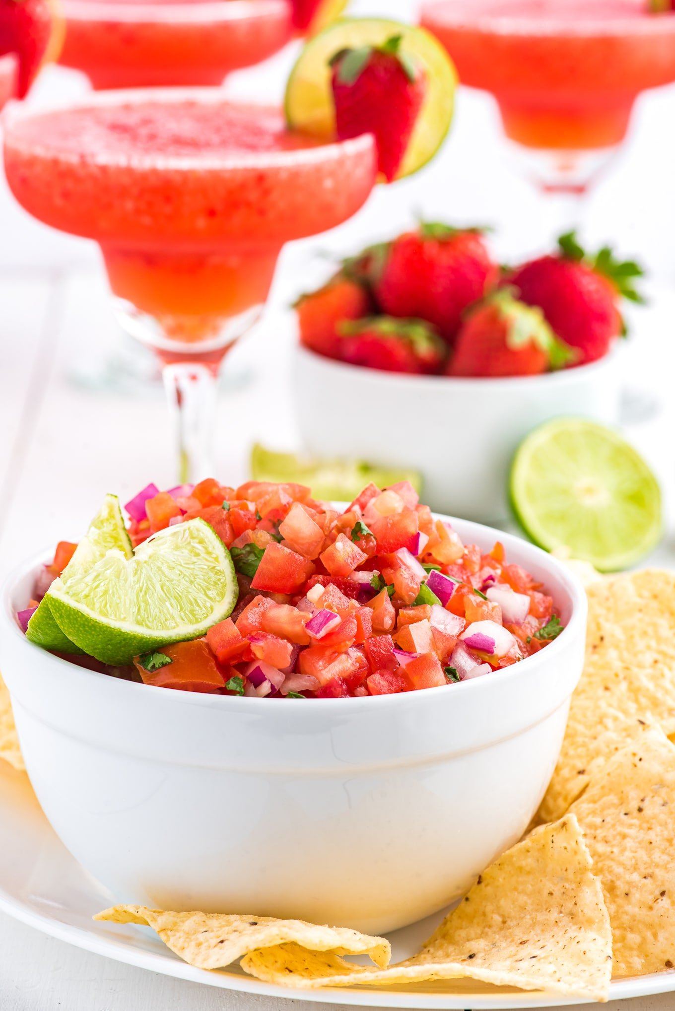 A bowl of pico de gallo with strawberry margaritas in the background.