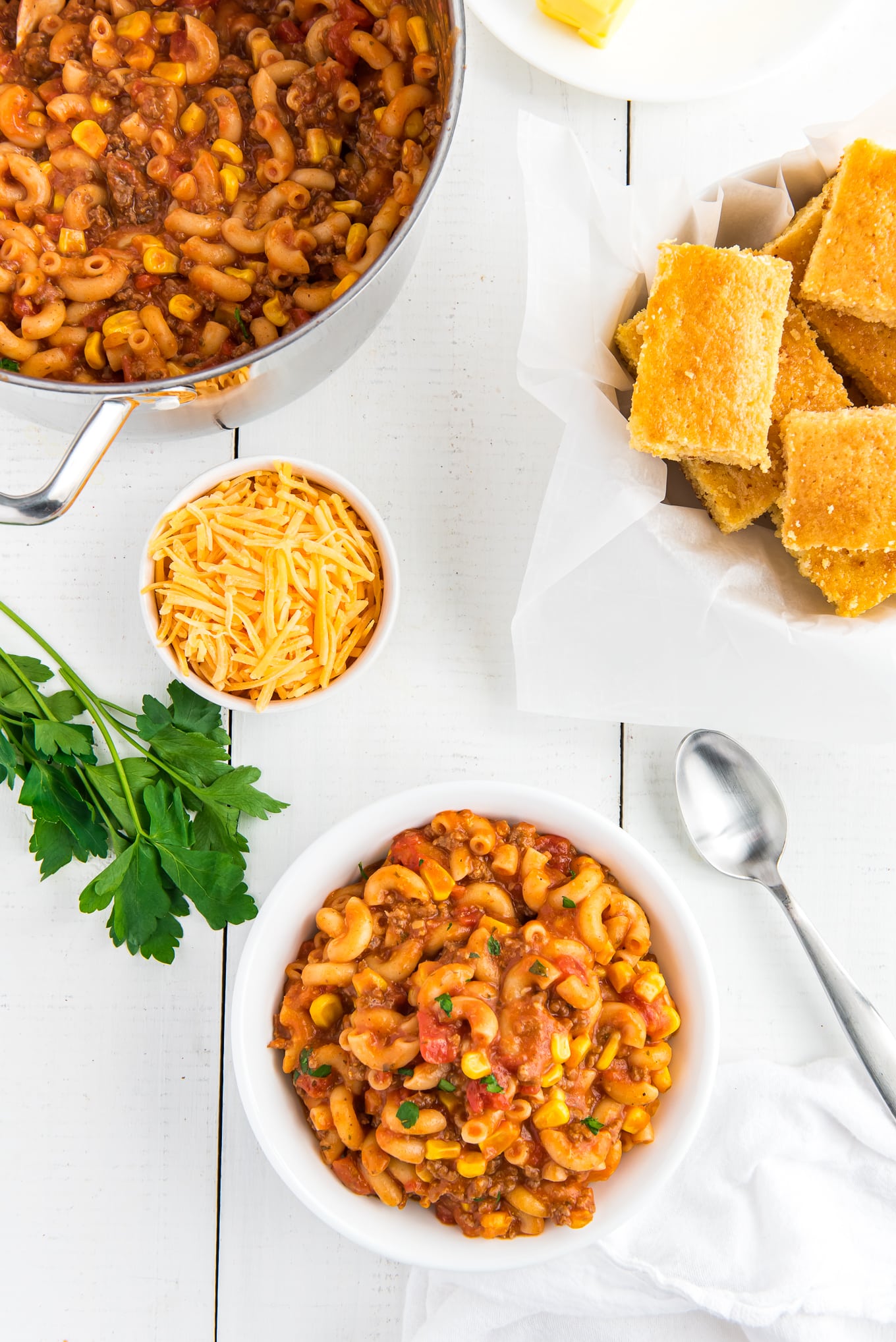 A bowl of beef goulash on the table with a bowl of shredded cheese, cornbread in a basket and a pot of more.