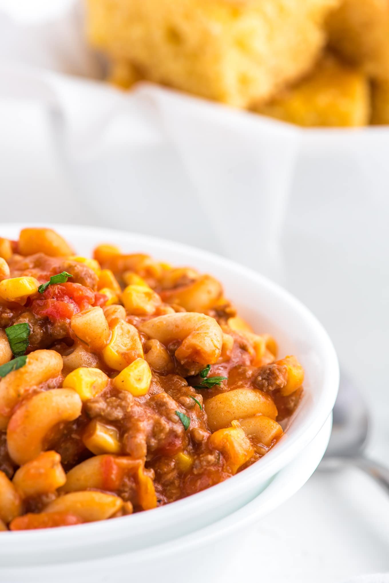 A side view of a bowl of American goulash with a basket of cornbread in the background.