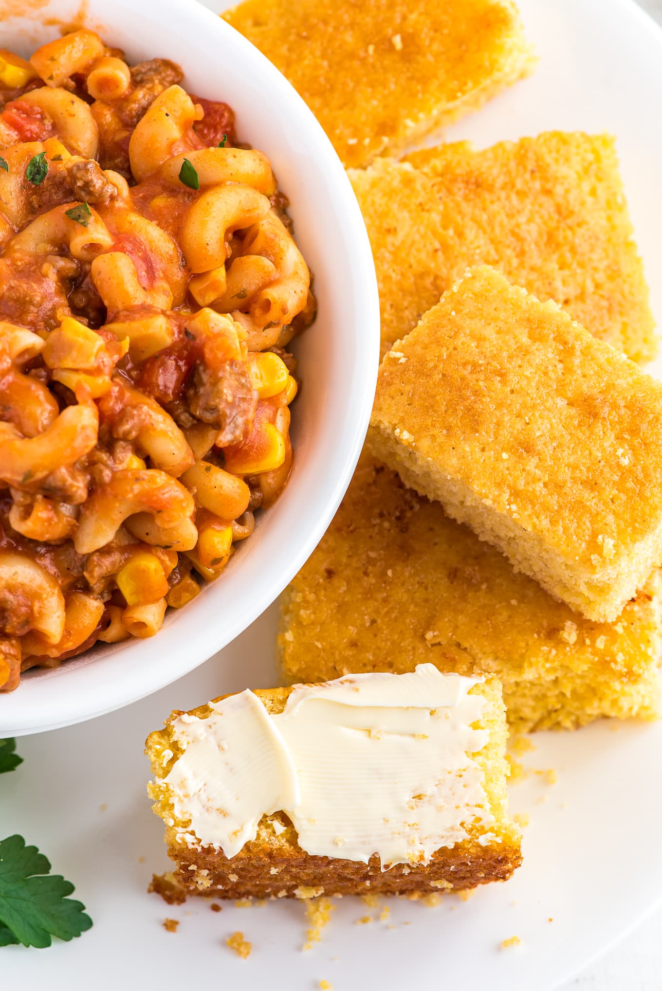 Jiffy cornbread squares on a plate with a bowl of goulash and butter on one piece.