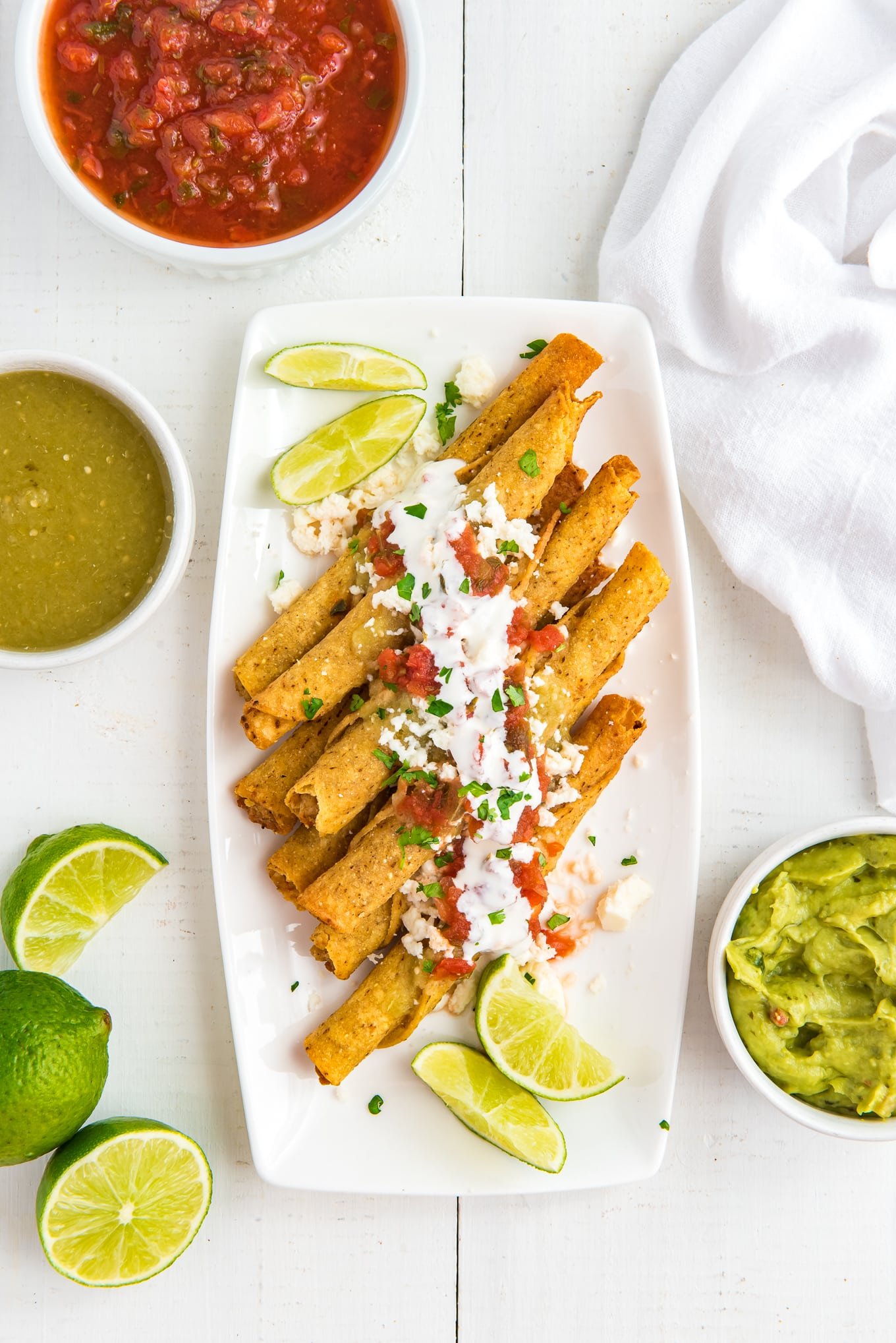 taquitos stacked on a plate topped with queso fresa cheese, salsa verde, Mexican sour cream, and salsa. And then put the guacamole and salsa on the side.