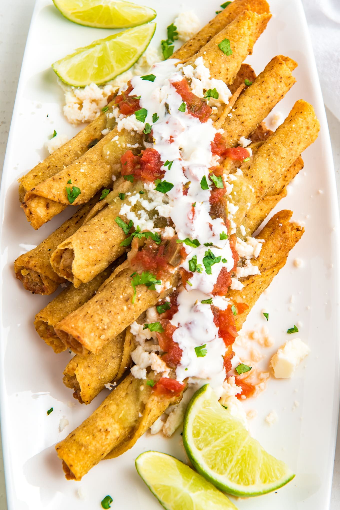 taquitos stacked on a plate topped with queso fresa cheese, salsa verde, Mexican sour cream, and salsa. 