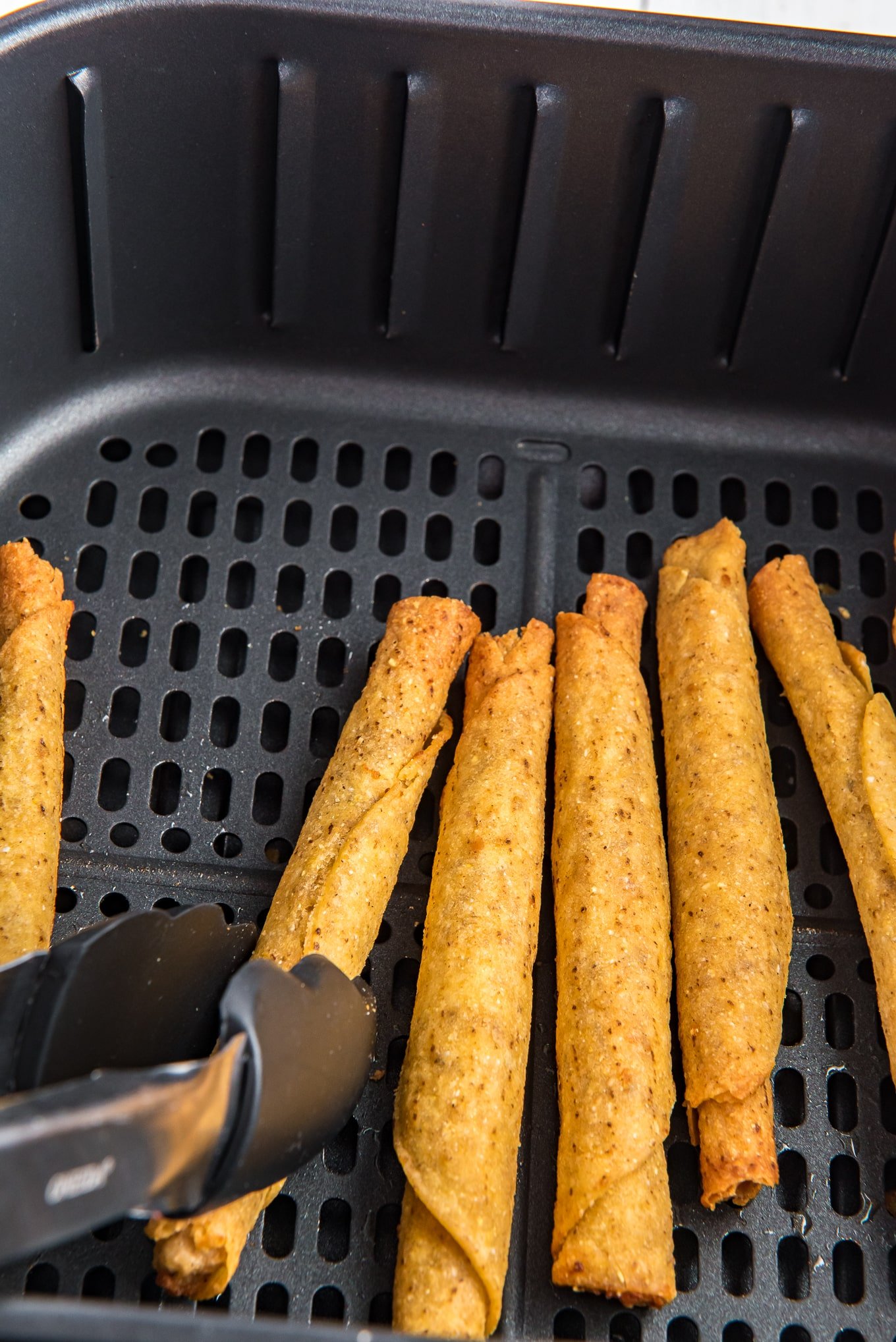 cooked taquitos in the air fryer basket being picked up with tongs. /