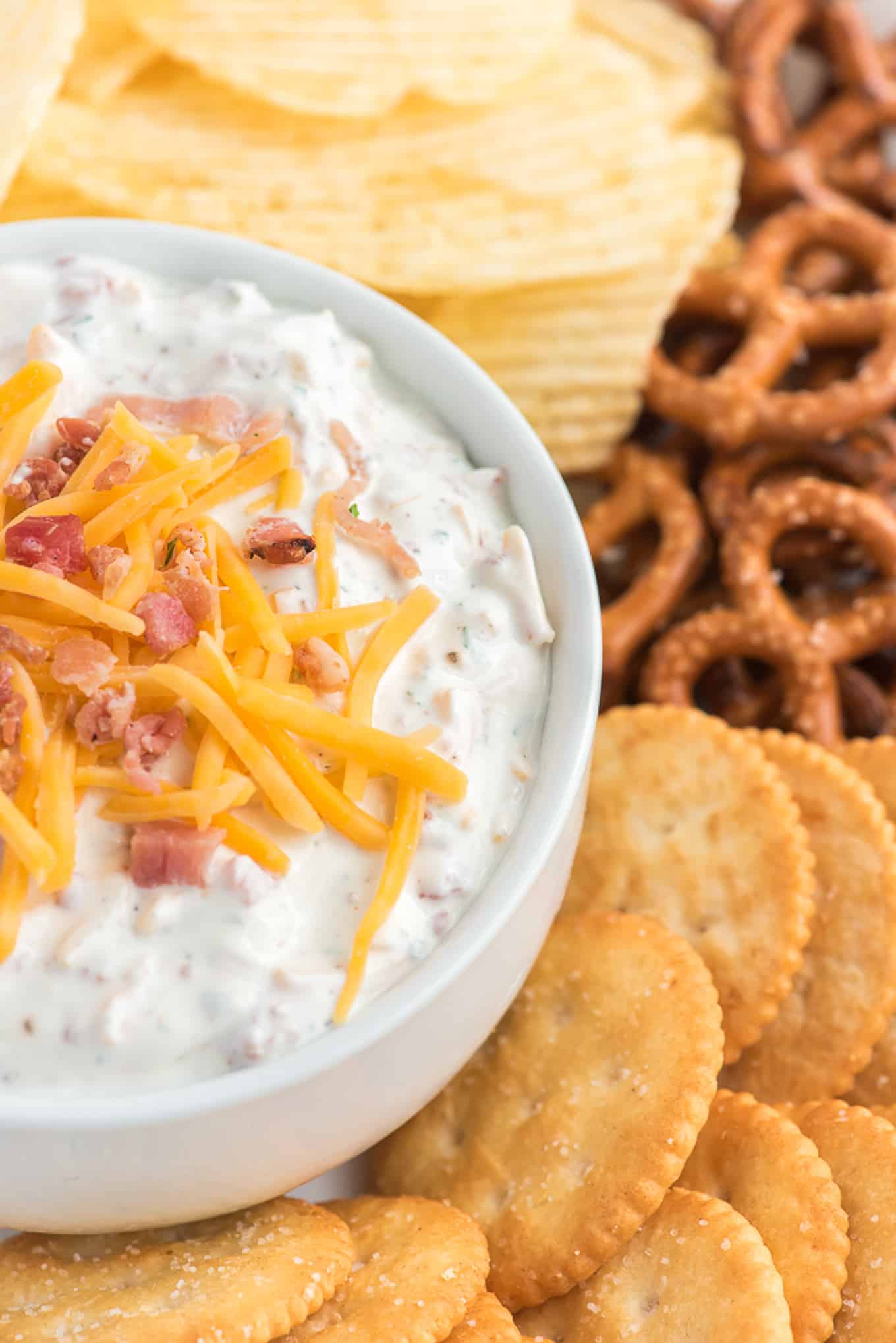 Crack dip topped with extra bacon and cheese surrounded by crackers, chips and pretzels. 