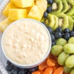 A white bowl of cream cheese pineapple dip on a plate with fruit.