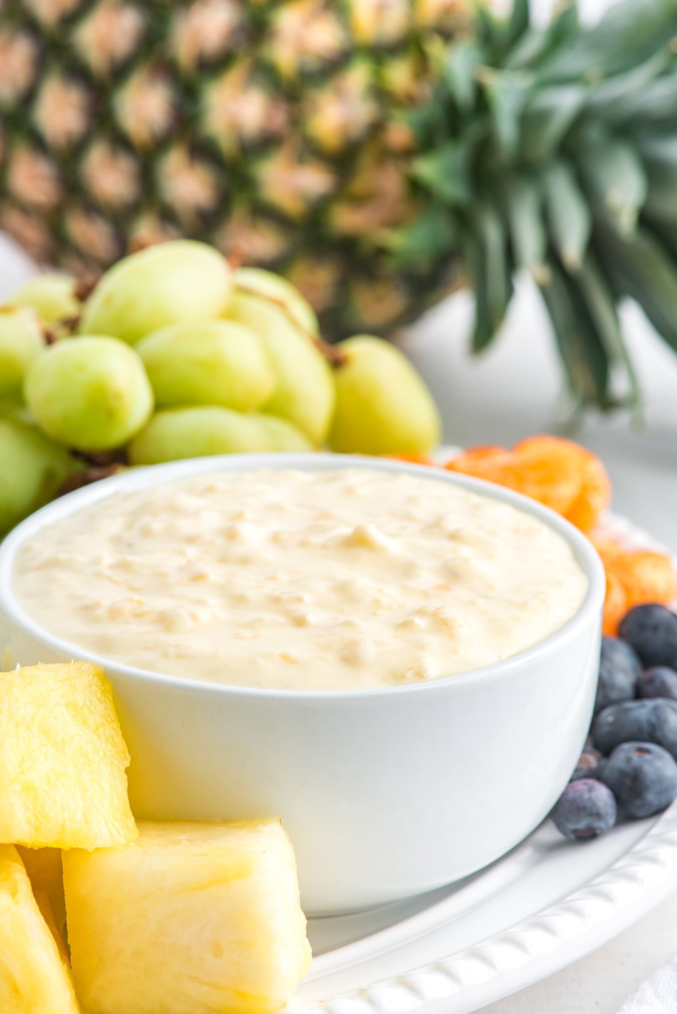 A side shot of a white bowl of cream cheese pineapple dip on a fruit plate with pineapple in the background.