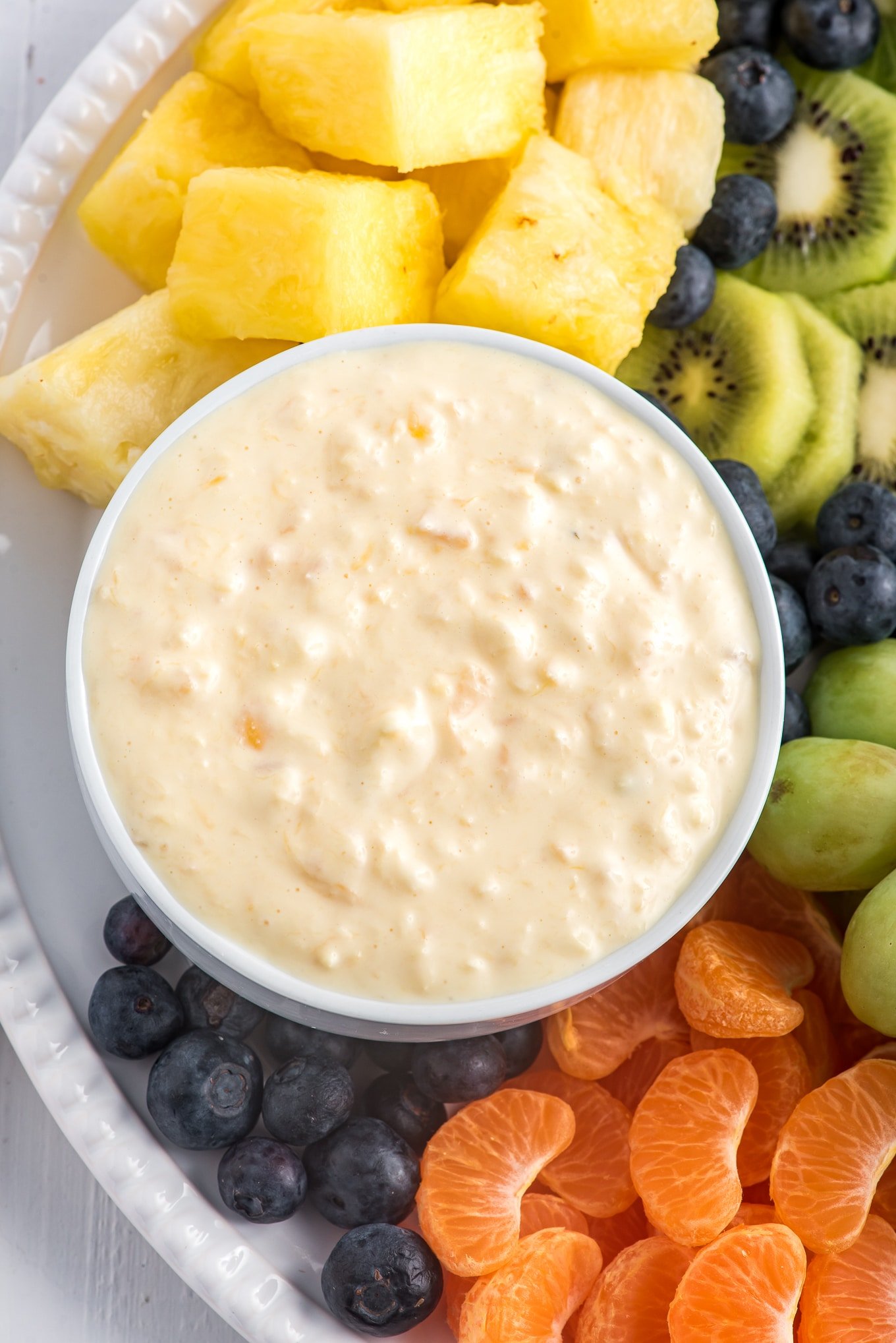 An overhead shot of a small white dish of cream cheese pineapple dip on a platter of fruit.