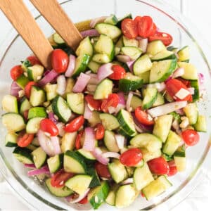 A bowl of cucumber tomato onion salad on the table with wooden servers in the bowl.