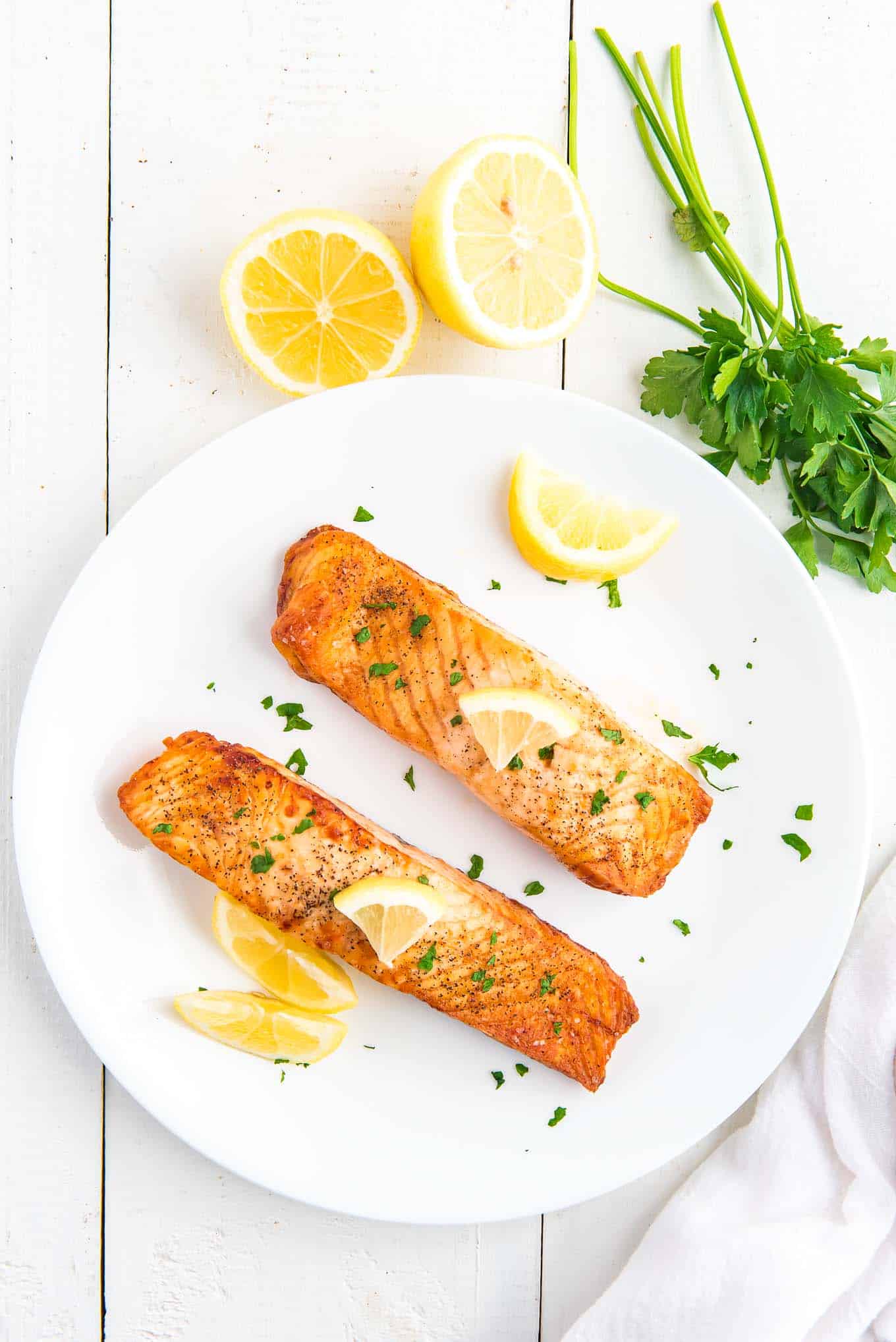 Two pieces of air fryer frozen salmon the table on a white plate with lemon wedges and parsley.