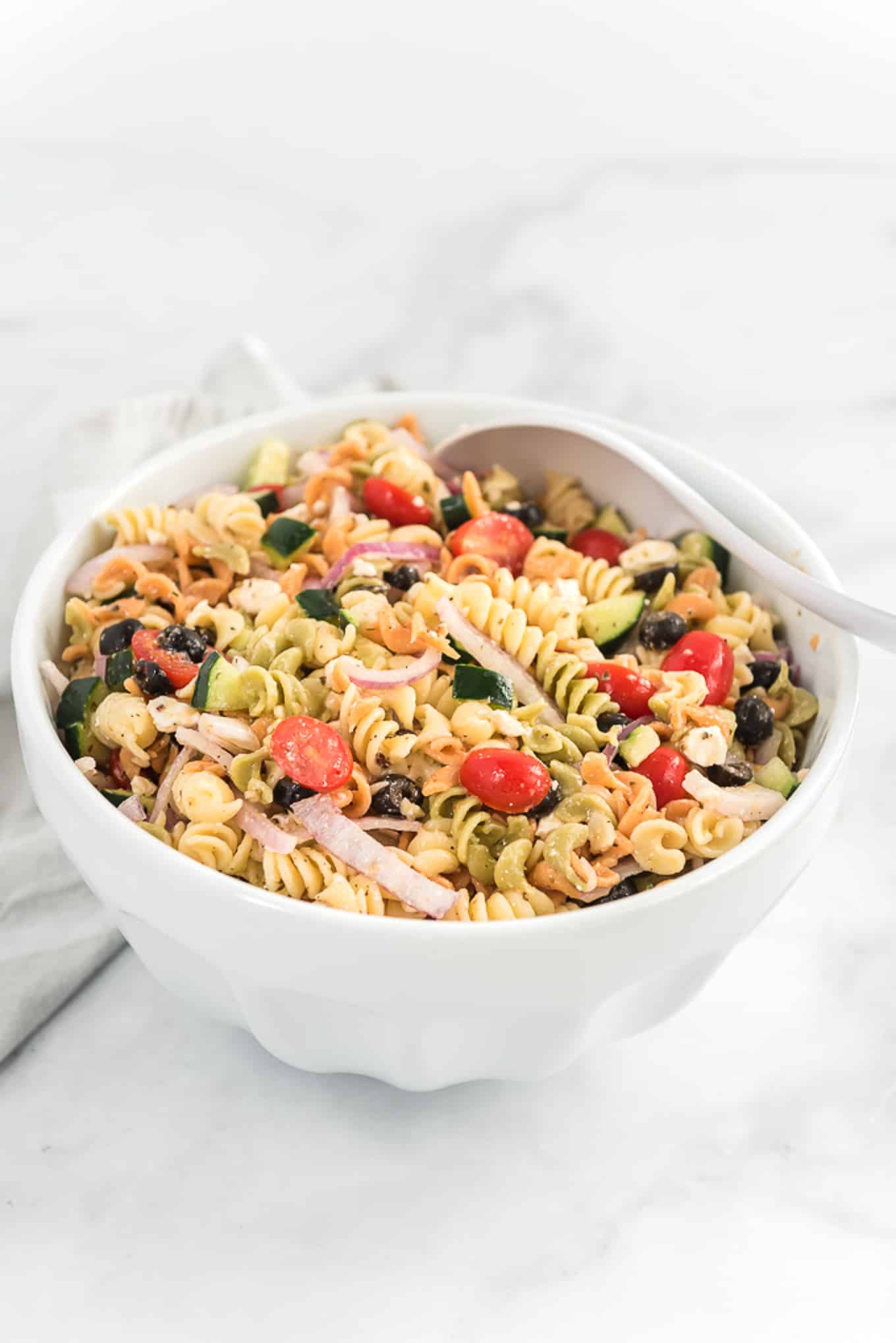 A white bowl of Greek pasta salad on the table with a white serving spoon in the bowl on its side.