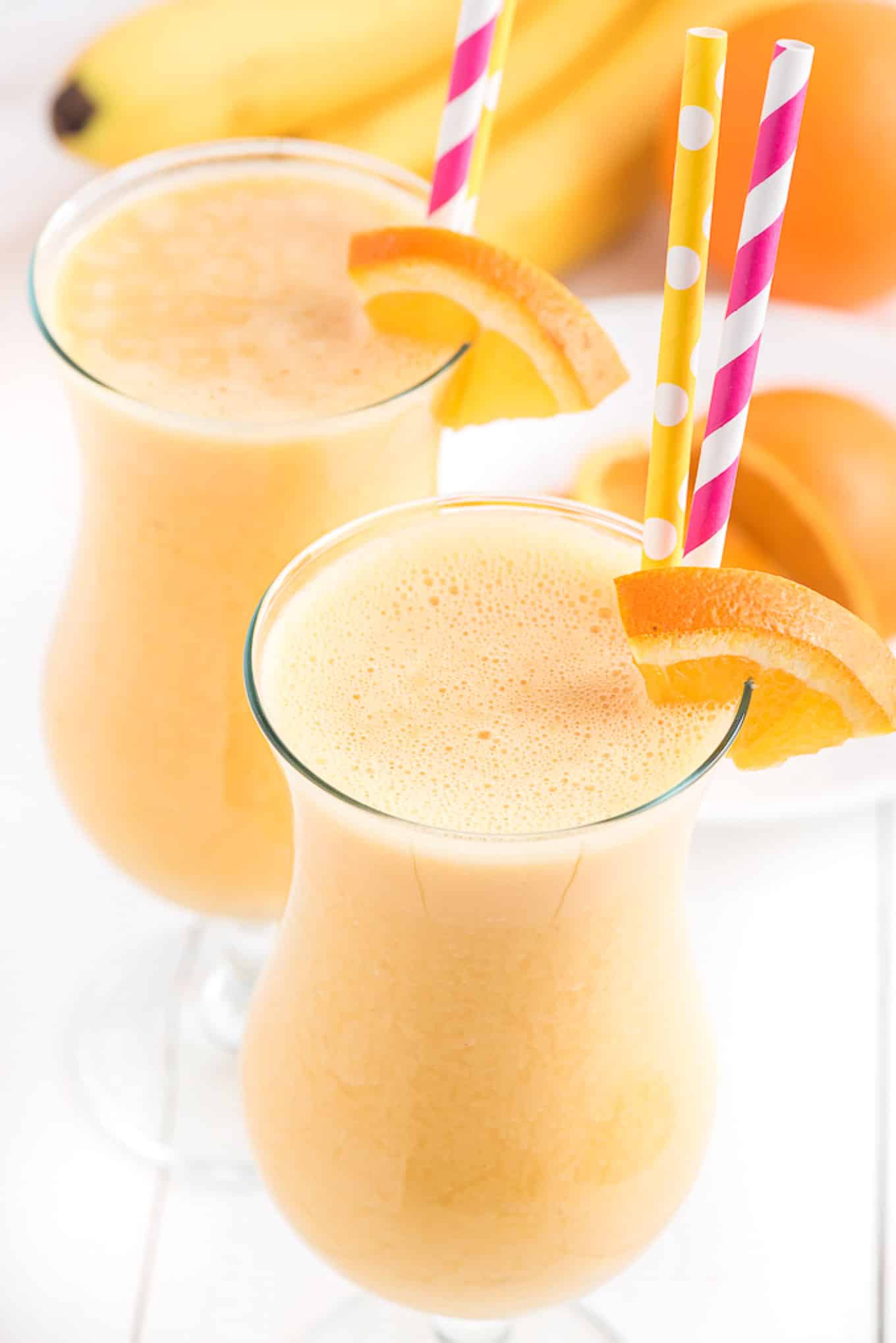 Tall glass of orange smoothie with an orange slice on the side and a pink straw. 