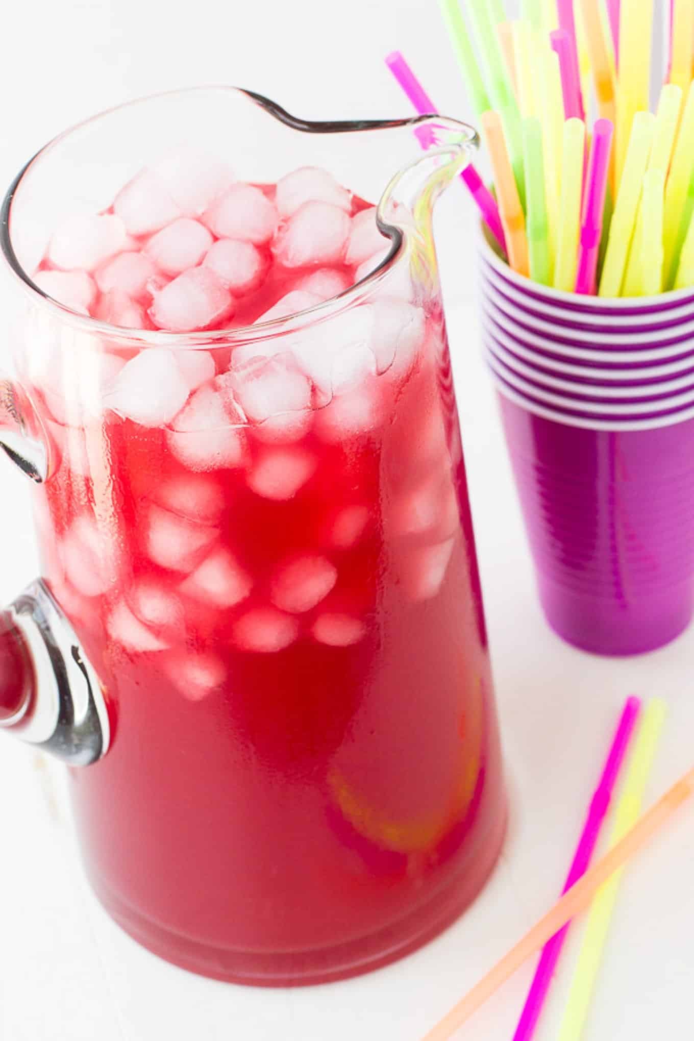 Tall pitcher of party punch and ice with solo cups behind it. 