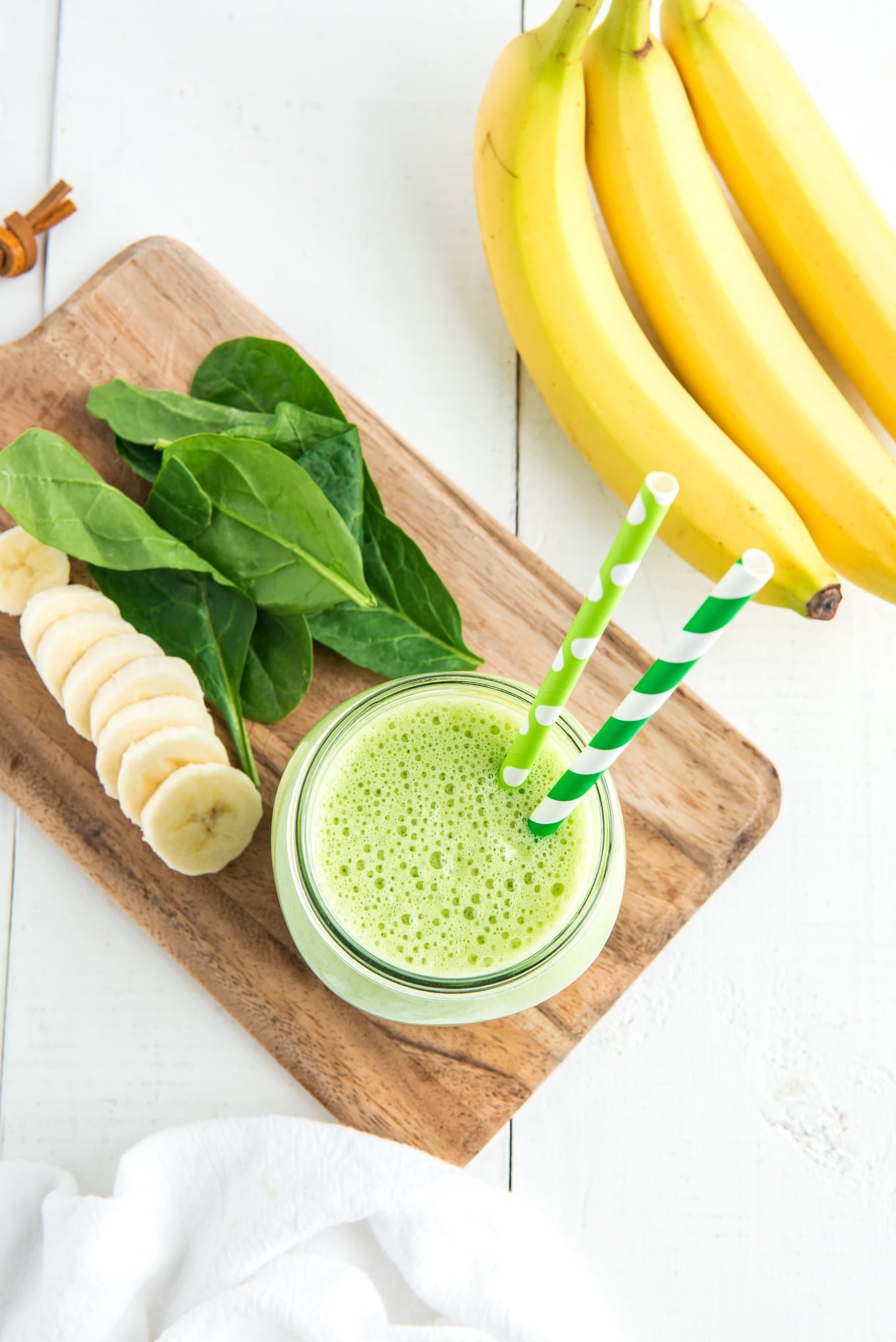 Spinach smoothie in a glass with two straws and some sliced bananas and fresh spinach leaves on the table next to the glass.
