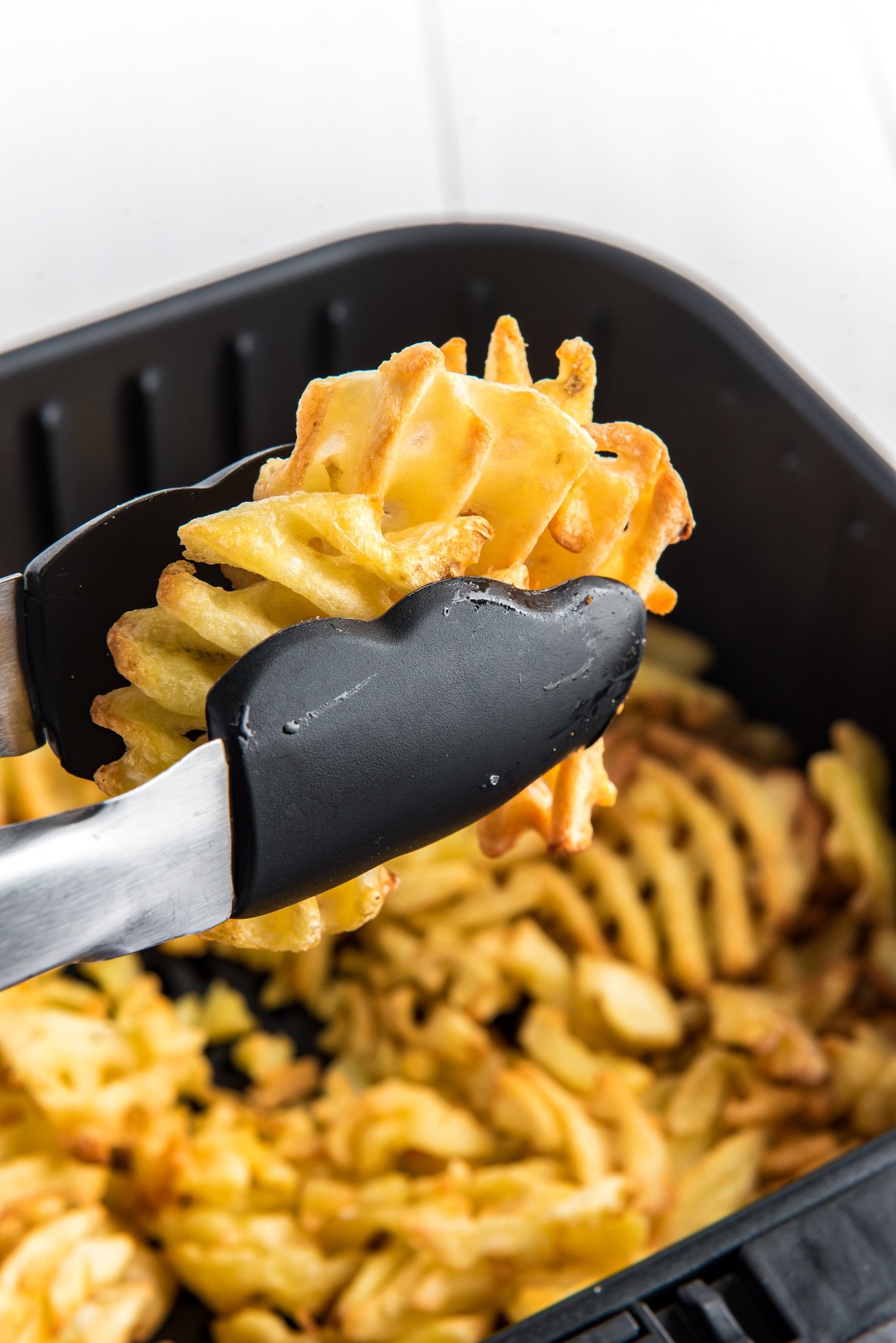 Taking the perfectly crisp waffle fries out of the air fryer with kitchen tongs. 