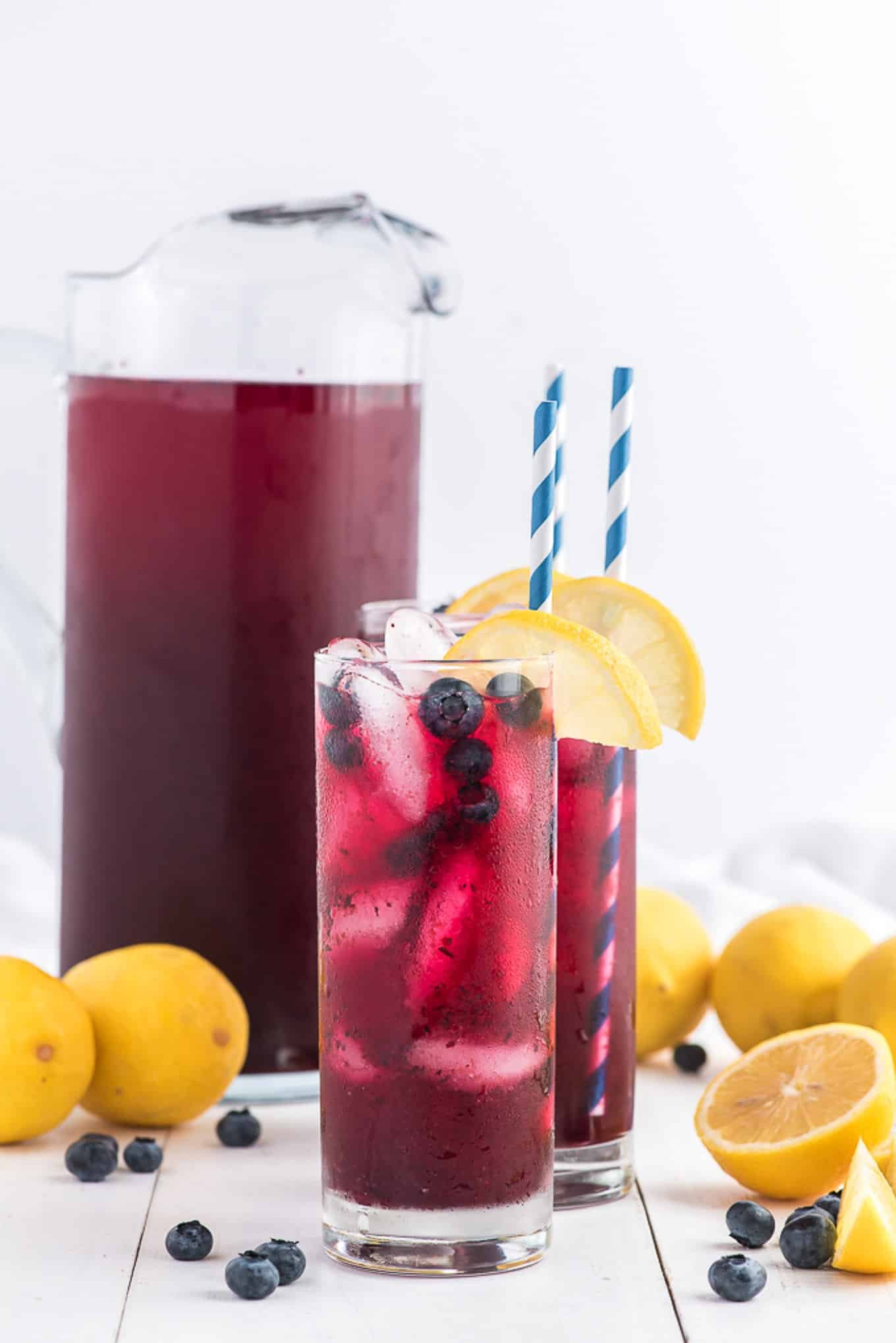 Two tall glasses of blueberry lemonade with lemon slices and straws surrounded by lemons and blueberries on the counter.