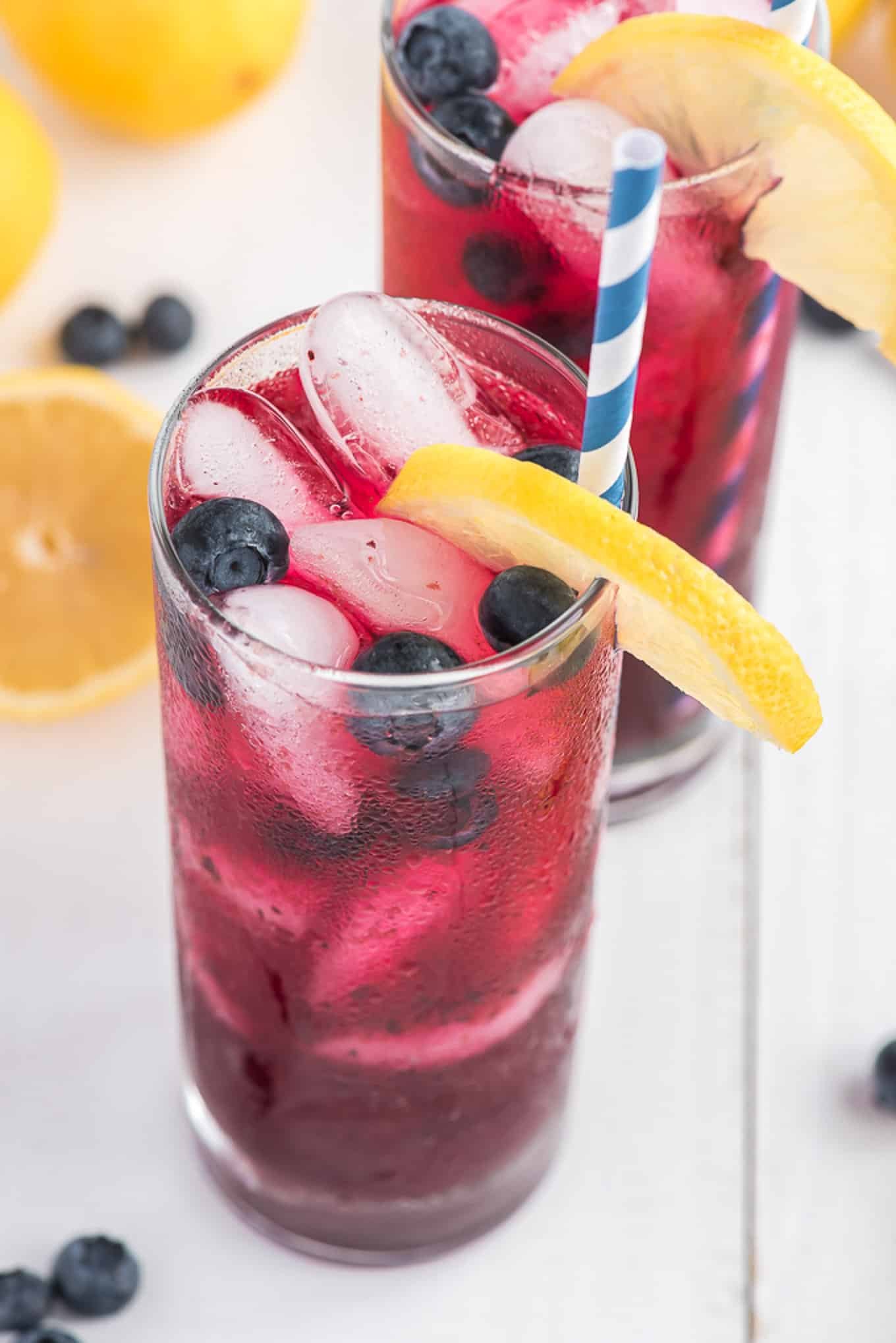 An angled shot of two tall glasses of blueberry lemonade with striped blue straws and lemon wedges.