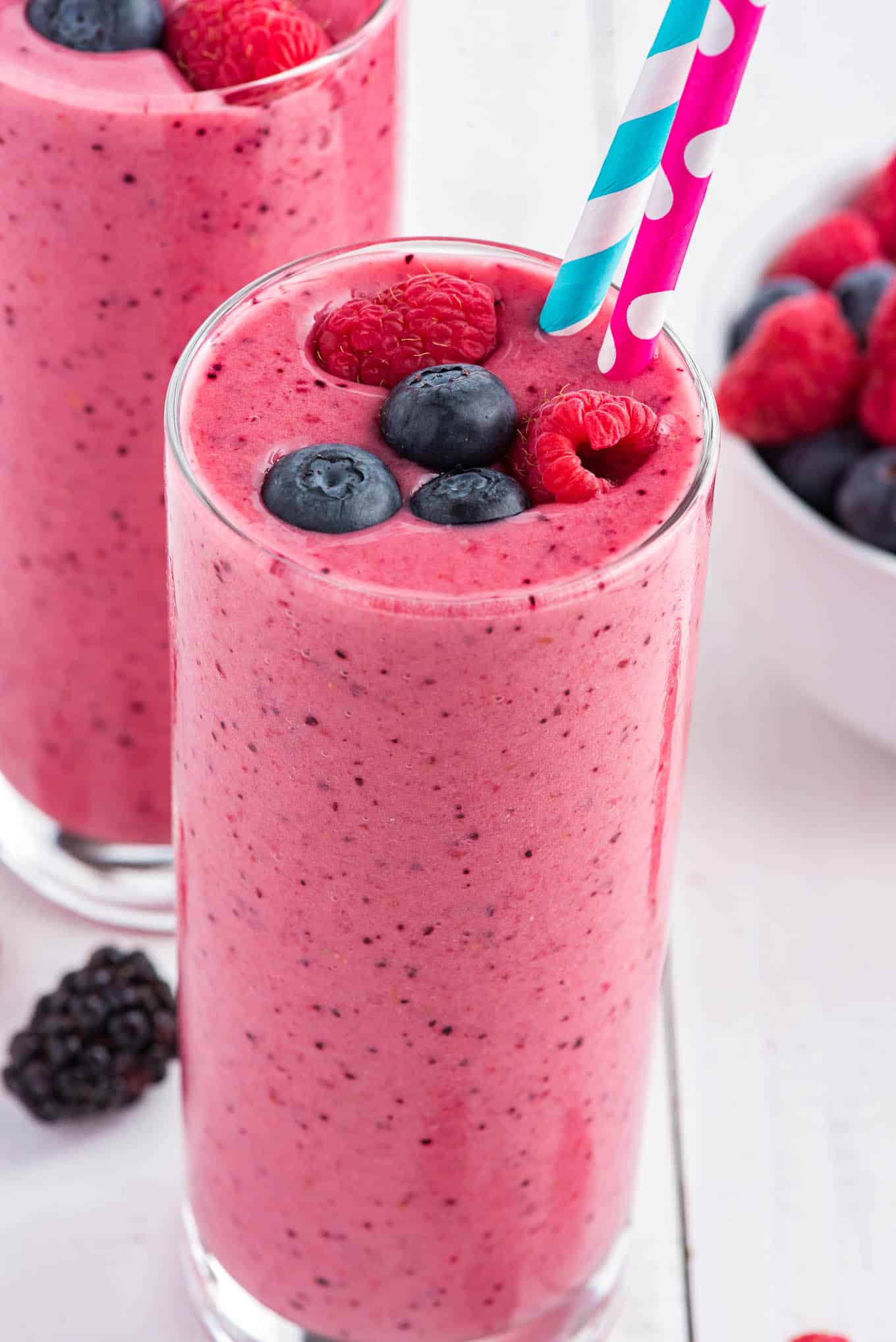 A glass of breakfast smoothie garnished with blueberries and raspberries on the table with another in the background and a bowl of berries.