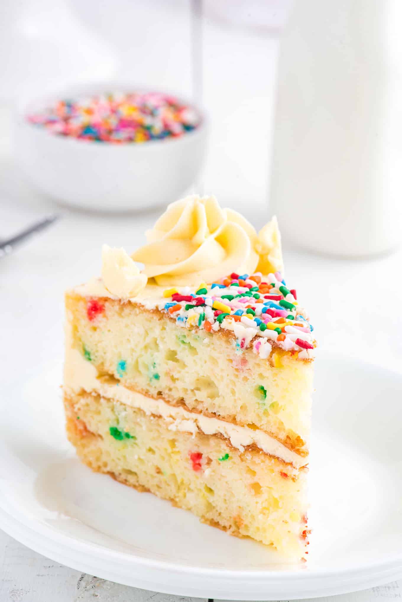 A slice of funfetti cake on a plate with a bowl of sprinkles and jar of milk in the background.
