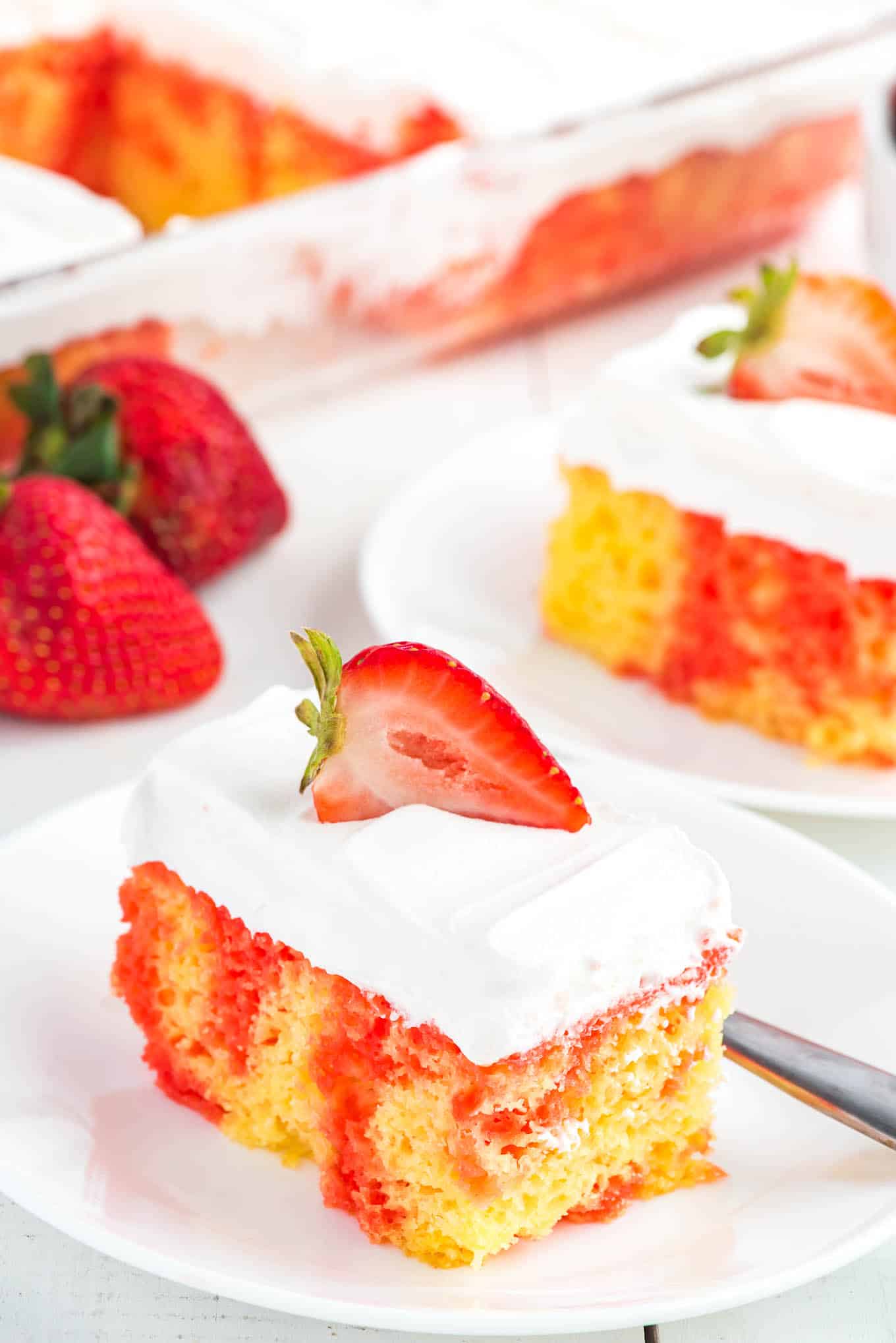 A square of jello poke cake topped with a strawberry half on a plate.